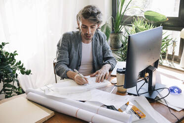 Confident male architect sketching blueprint on paper at desk in home office - MASF43342