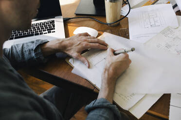 Midsection of male architect sketching blueprint on paper at desk in home office - MASF43343