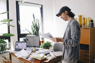 Side view of male architect analyzing blueprint while standing by desk in home office - MASF43349