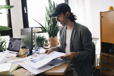 Male architect analyzing blueprint while sitting at desk in home office - MASF43351