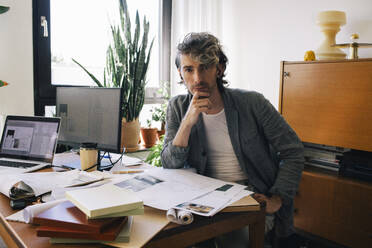 Portrait of male architect sitting with hand on chin at desk in home office - MASF43353