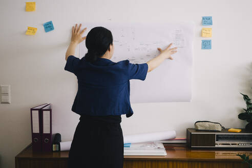 Rear view of female architect examining blueprint on wall at home office - MASF43378