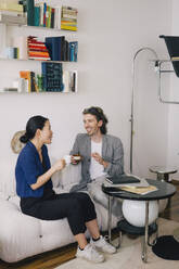 Full length of happy male and female architects with coffee cups sitting on sofa at home office - MASF43384
