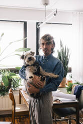 Portrait of male architect carrying dog standing at home office - MASF43389