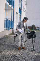 Full length of male architect holding blueprint rolls standing by bicycle on footpath - MASF43391