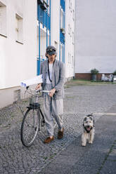 Full length of male architect holding blueprint rolls walking by bicycle and dog on footpath - MASF43392