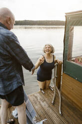 Happy senior woman in swimwear climbing houseboat by man - MASF43393