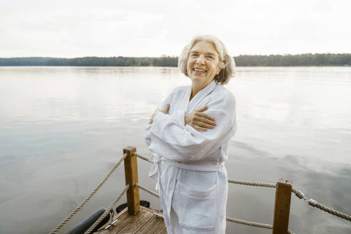 Portrait of happy senior woman wearing white bathrobe hugging self on houseboat near river against sky - MASF43395