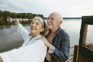 Senior man with hands on happy woman's shoulder on houseboat near river - MASF43396