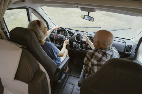 Senior man sharing smart phone with woman driving camper van during road trip - MASF43397