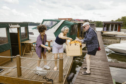 Full length of man giving basket to woman on houseboat against sky - MASF43442