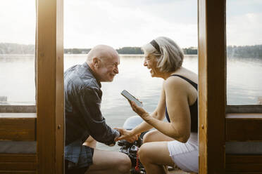 Happy senior couple with smart phone sitting on houseboat - MASF43452
