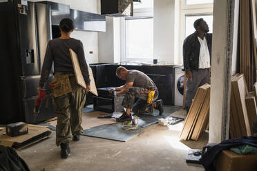 Female carpenter carrying plank while coworkers working in kitchen at site - MASF43468