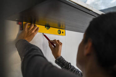 Female carpenter using bubble level and marking on wall with pencil - MASF43472