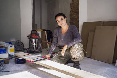 Portrait of confident female carpenter holding handsaw crouching in front of plank in apartment - MASF43498