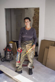 Portrait of confident female carpenter holding handsaw and ruler standing in apartment - MASF43499