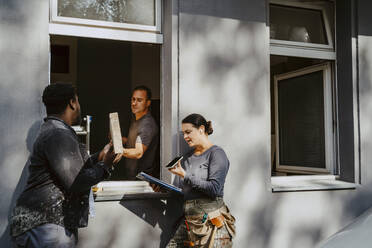 Workers passing plank through window while female construction manager talking on phone outside house - MASF43511