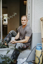 Portrait of smiling male carpenter holding coffee cup at doorway of house - MASF43518