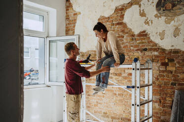 Man talking with woman sitting on ladder in front of brick wall during home renovation - MASF43531