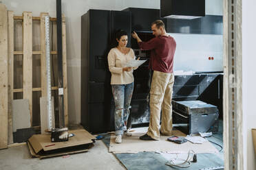 Couple discussing over document while fixing cabinet together in kitchen during home renovation - MASF43535