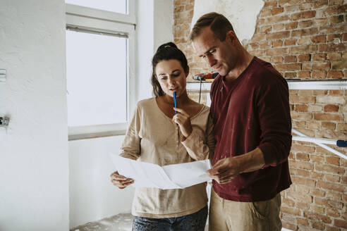 Couple discussing over blueprint in front of brick wall while renovating home - MASF43546