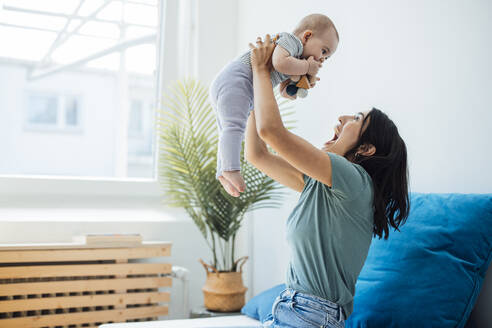 Cheerful mother lifting daughter sitting on bed at home - JOSEF23856