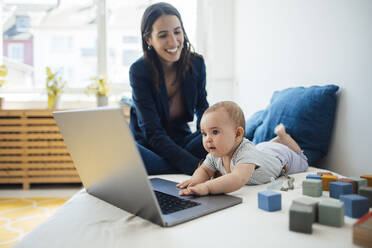 Happy businesswoman sitting by baby girl playing with laptop on bed - JOSEF23869