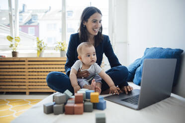 Lächelnde Geschäftsfrau, die mit einem kleinen Mädchen auf einem Bett zu Hause sitzt und einen Laptop benutzt - JOSEF23870