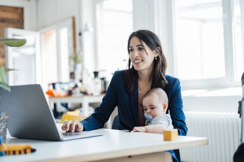 Smiling businesswoman sitting with baby girl using laptop at home - JOSEF23878
