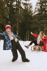 Cheerful boy pulling mother sitting on sled in winter forest - MCHF00012