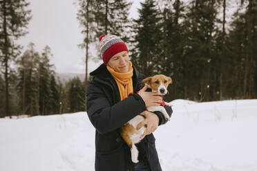 Smiling man holding pet dog in winter forest - MCHF00016