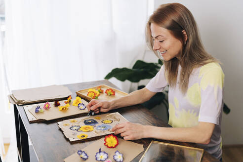 Smiling woman arranging dried flowers on cardboard at home - TILF00075