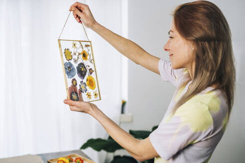 Woman holding transparent frame decorated with dried flowers at home - TILF00077