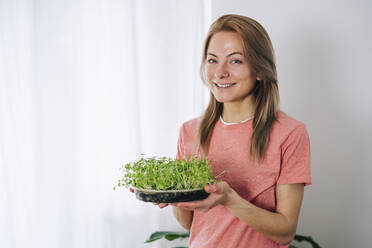 Smiling woman holding broccoli microgreen at home - TILF00083
