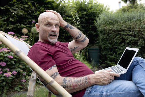 Surprised freelancer sitting with laptop on deck chair in garden - FLLF00909