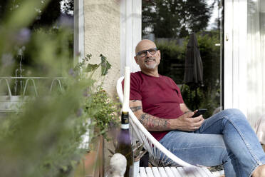 Lächelnder Mann sitzt mit Smartphone vor einem Haus - FLLF00913