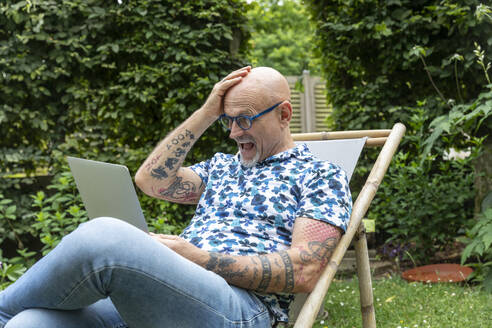 Überrumpelter Mann mit Kopf in der Hand, der im Garten auf einen Laptop schaut - FLLF00920