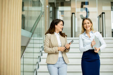 Two cute young business women walking on stairs in the office hallway - INGF13105