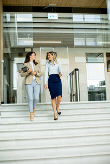 Two cute young business women walking on stairs in the office hallway - INGF13106