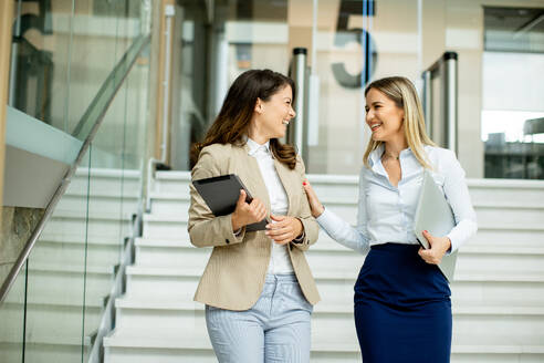 Two cute young business women walking on stairs in the office hallway - INGF13107