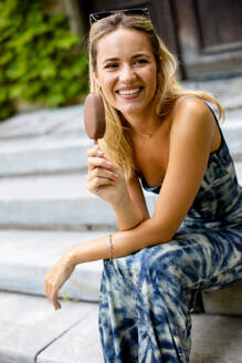 Pretty young woman sitting on stairs and eating ice cream on stick - INGF13111