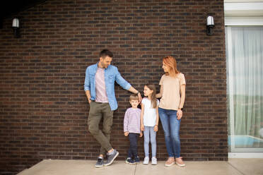 Family with a mother, father, son and daughter standing by the wall of a brick house - INGF13132