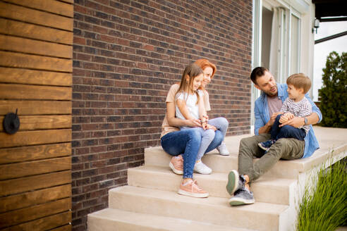 Family with a mother, father, son and daughter sitting outside on steps of a front porch of a brick house - INGF13135