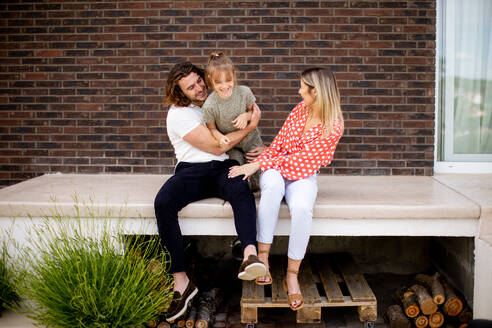 Family with a mother, father and daughter sitting outside on steps of a front porch of a brick house - INGF13138