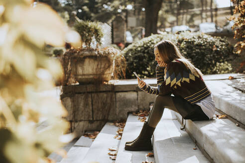 Pretty young woman sitting on a stairs with mobile phone in autumn park - INGF13147