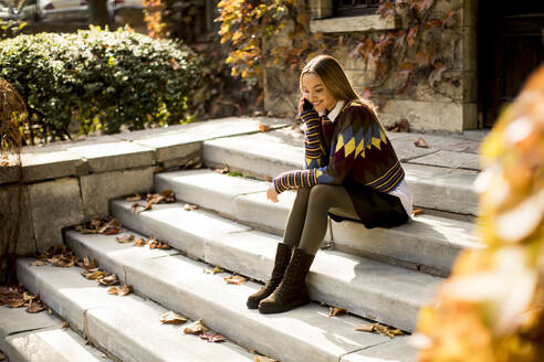 Pretty young woman sitting on a stairs with mobile phone in autumn park - INGF13150