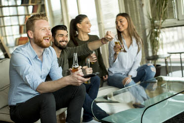 Group of young friends looking at the TV, drinking cider and having fun in the room - INGF13158