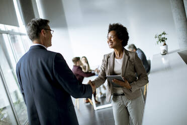 African American businesswoman holding digital tablet and looking at handsome colleague while shaking hands in office - INGF13168