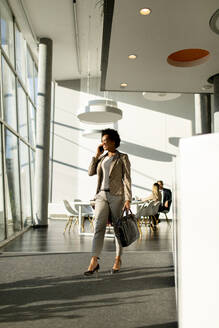 African American business woman with bag using mobile phone and walking in the office - INGF13181