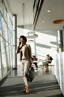 African American business woman with bag using mobile phone and walking in the office - INGF13182